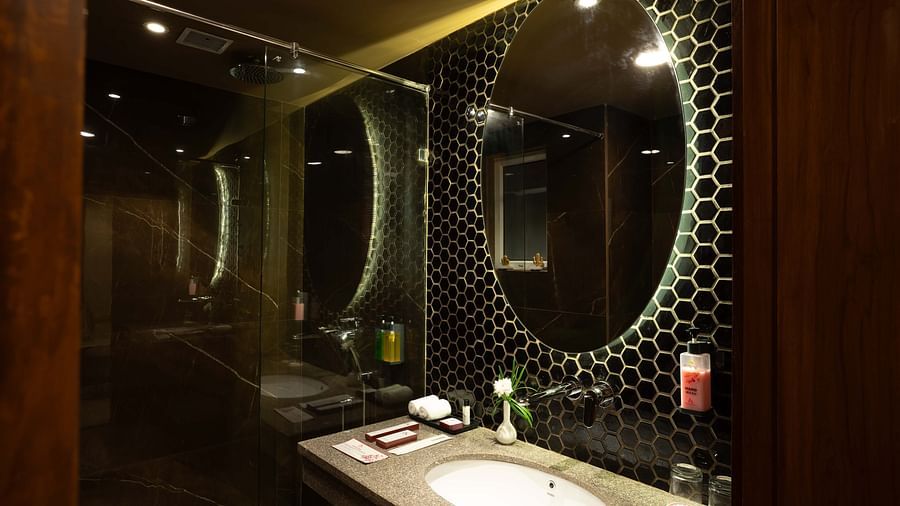 Round shaped mirror above the washbasin with transparent glass divider separating the showering and toilet areas in the Deluxe room's bathroom at Inde Signature Jaipur.