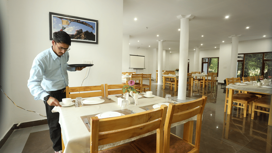 A dining space with chairs, plates placed on tables, a person serving in a cup with saucer, and wall arts at Indus Serinity, Leh Ladakh.