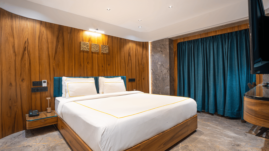 A bedroom with white bedding, wooden interiors and blue curtains at Infiniti Hotel, Indore.