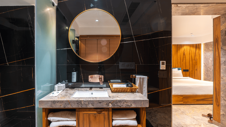 A washbasin with circular mirror, black tiled wall and toiletries at Infiniti Hotel, Indore.