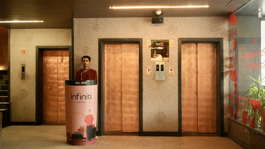 An elevator lobby at Infiniti Hotel, Indore featuring 3 lift doors, staff member at a cylindrical desk, and wall lights.