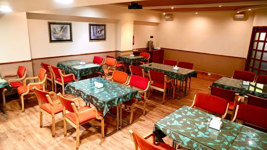 An empty dining room with multiple tables, patterned tablecloths, and several upholstered chairs at Infiniti Hotel, Indore.