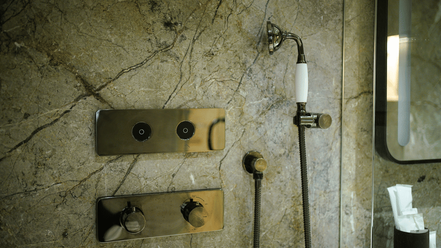 A bathroom at Infiniti Hotel, Indore featuring wall shower head, handheld shower, tap controls, and marble tiled wall.