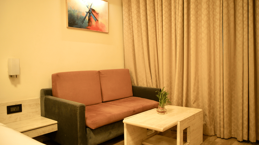 A bedroom seating corner at Infiniti Hotel, Indore featuring sofa, coffee table, plant, and window curtains.