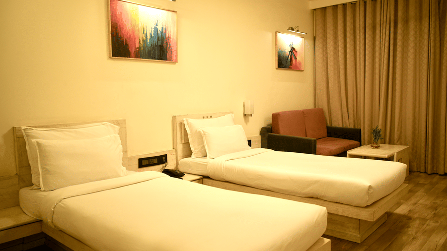 A bedroom at Infiniti Hotel, Indore featuring 2 single beds, sofa, curtains, and wall art above headboards.