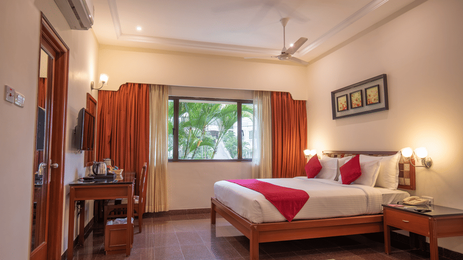 A well-lit Junior Suite features a king size bed, bedside tables with lamps, a window with curtains, a ceiling fan, a wall-mounted TV, and a small desk with a chair