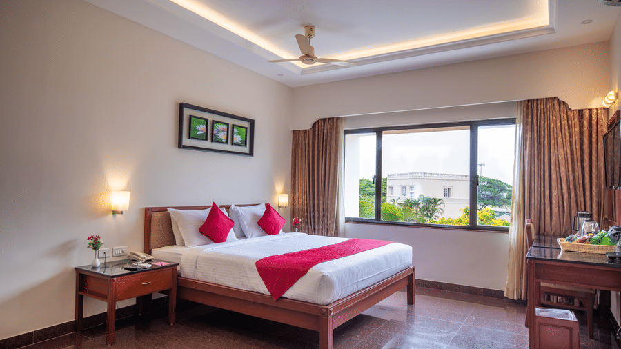 A bright Executive Room with a king sized bed, pillows, bedside tables with lamps, a desk with a chair, curtains framing a large window, wall art, and a ceiling fan