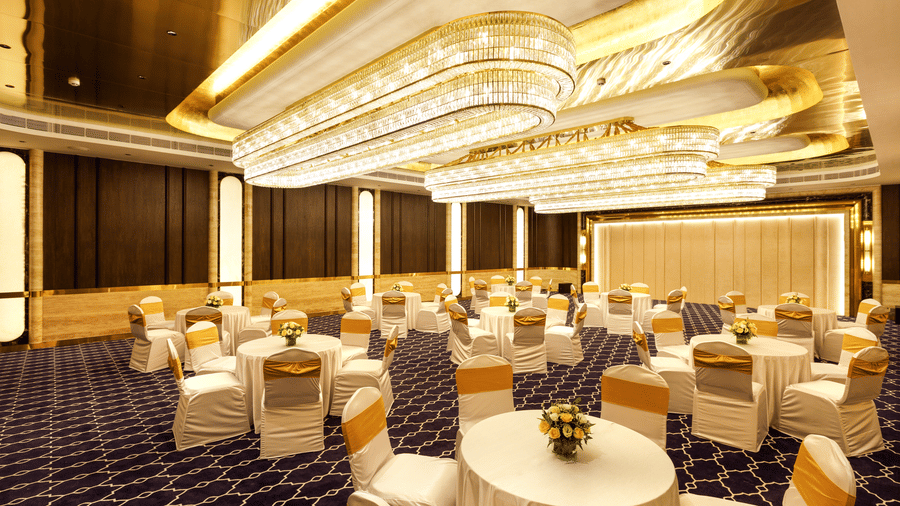 Luxurious banquet hall set up with round tables and white chairs with gold sashes, under elegant chandeliers, at Jenneys Residency, Coimbatore.