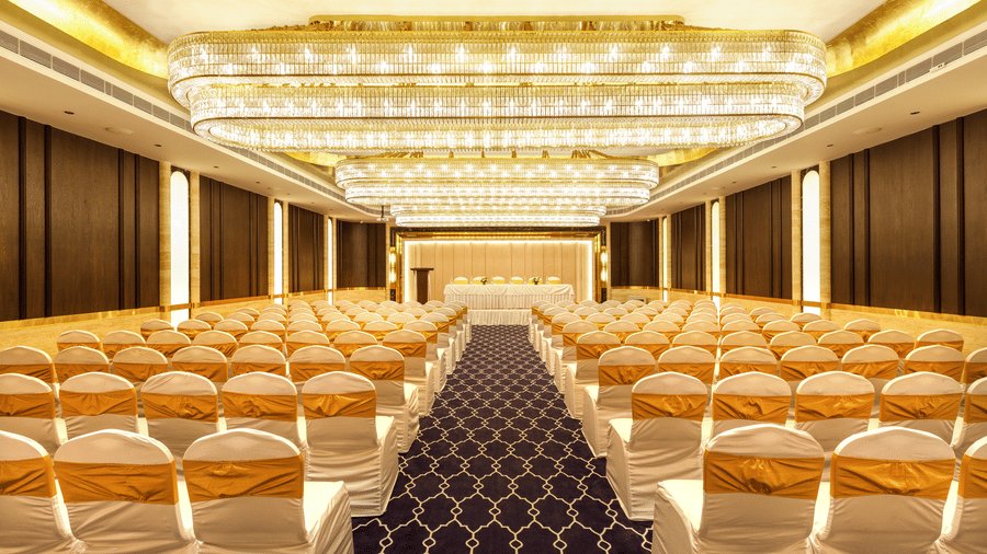 Elegant banquet hall with rows of white and gold-covered chairs facing a stage, illuminated by chandeliers, at Jenneys Residency, Coimbatore.