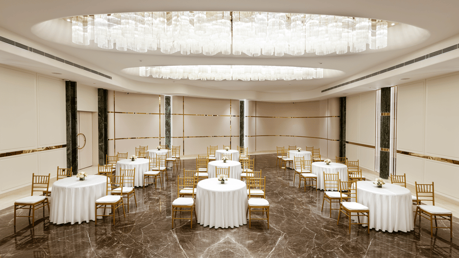 Emerald Hall set up with round tables and gold chairs beneath elegant chandeliers at Jenney Residency, ideal for celebrations.