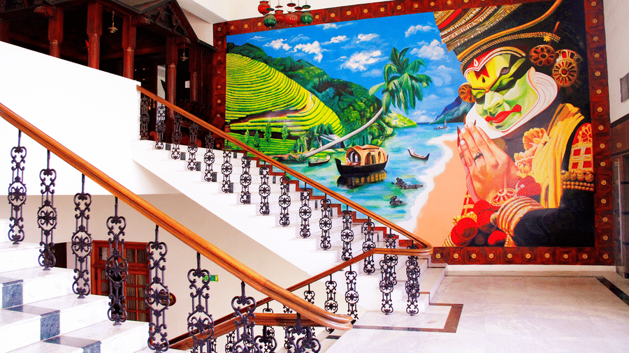 Staircase with ornate railing beside a vibrant Kathakali mural and tropical scenery - Jenneys Residency, Coimbatore