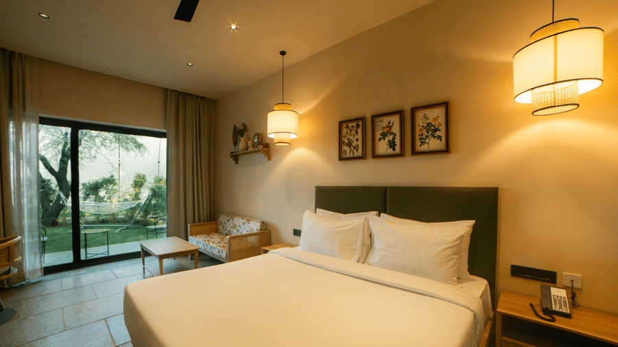 A king size bed sits under 3 framed pictures, with a sliding glass window opening to an outdoor lawn and hanging lamps at Jivana Wellness Retreat, Bhopal.