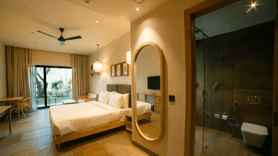 A room with a king size bed with white bedding, a large vertical oval mirror on the wall, and a window overlooking the greens at Jivana Wellness Retreat, Bhopal.