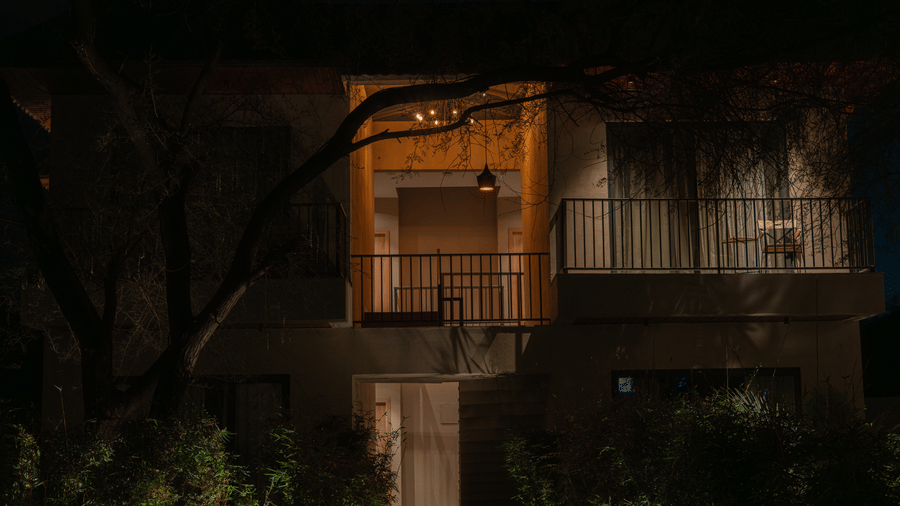 The exterior of a building at night features glowing windows, metal railings on balconies, and dark silhouettes of trees and shrubbery at Jivana Wellness Retreat, Bhopal.