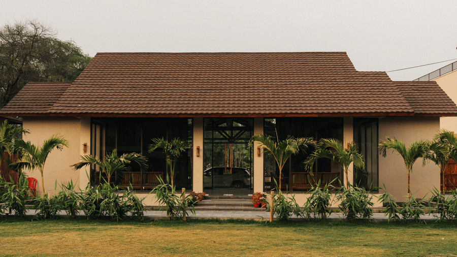 A sloped roof building with glass windows is surrounded by small palm trees and a flat grass lawn at Jivana Wellness Retreat, Bhopal.