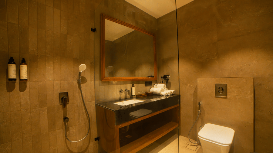 A bathroom view showing a wooden vanity with a white sink, toilet, grey tiled walls, and a shower head with tiered lighting at Jivana Wellness Retreat, Bhopal.
