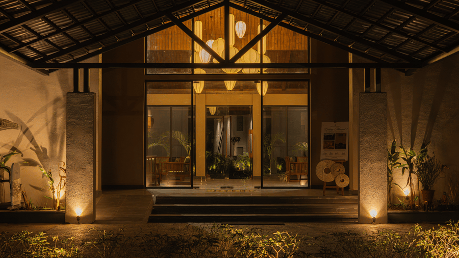 A building entrance with large glass doors is illuminated by floor spotlights, featuring a wide overhanging roof and stone pillars at Jivana Wellness Retreat, Bhopal.