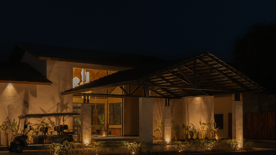 A nighttime shot of the entrance showing warm lighting reflecting off the glass and stone, under a large triangular wooden roof structure at Jivana Wellness Retreat, Bhopal.