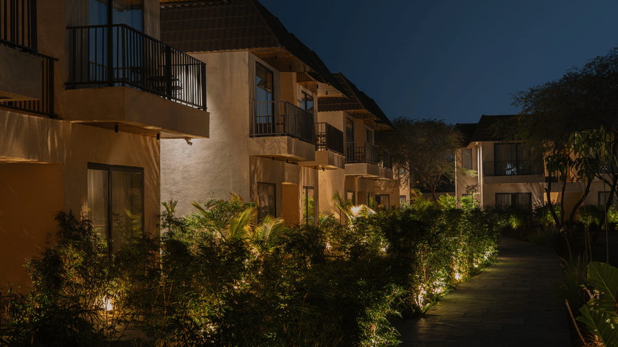 A stone path at night is lined with thick green bushes and warm ground lights, leading toward a row of 2-storey buildings with balconies at Jivana Wellness Retreat, Bhopal.