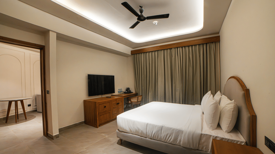 A suite with a king size bed, a television on a wooden stand, a ceiling fan, and long curtains covering a window at Jivana Wellness Retreat, Bhopal.