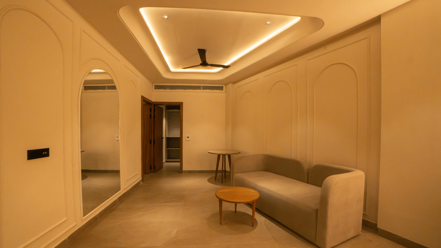 An indoor living area featuring a tan sofa, a small round wooden coffee table, recessed ceiling lighting, and a large wall mirror at Jivana Wellness Retreat, Bhopal.