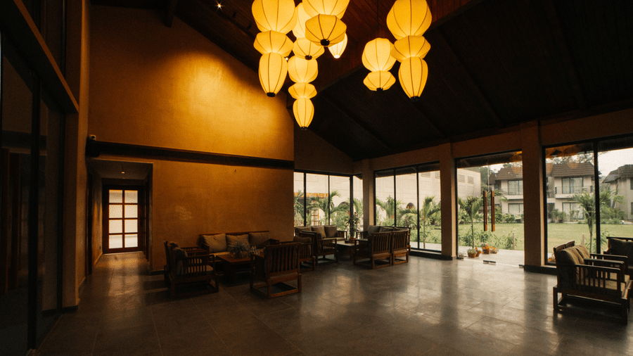 A dark lobby featuring several large hanging paper lanterns, lounge seating, and floor-to-ceiling windows looking out to a garden at Jivana Wellness Retreat, Bhopal.
