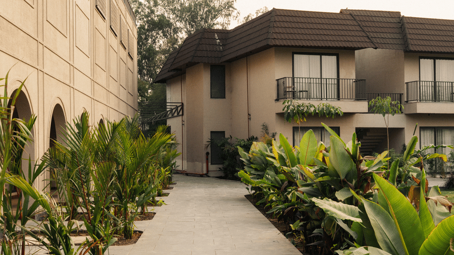 A paved path runs between a tan building and lush green plants leading toward a building at Jivana Wellness Retreat, Bhopal.