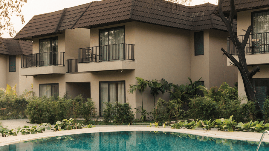 A view of a 2-storey building with dark roofs and metal balconies facing a blue swimming pool and tropical landscape at Jivana Wellness Retreat, Bhopal.