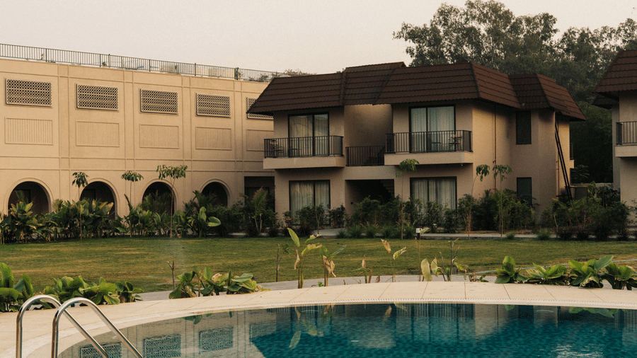 A curved swimming pool sits in front of a 2-storey tan building with balconies and a green lawn under an overcast sky at Jivana Wellness Retreat, Bhopal.