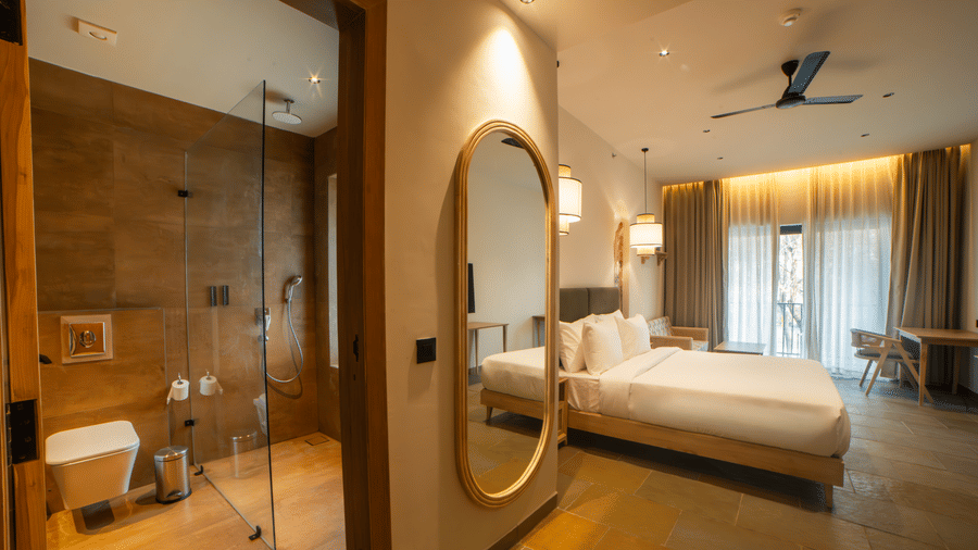 A view of the room from the doorway showcasing a full-length oval mirror, a white bed, a desk area, and a glimpse into a tiled bathroom on the left at Jivana Wellness Retreat, Bhopal.