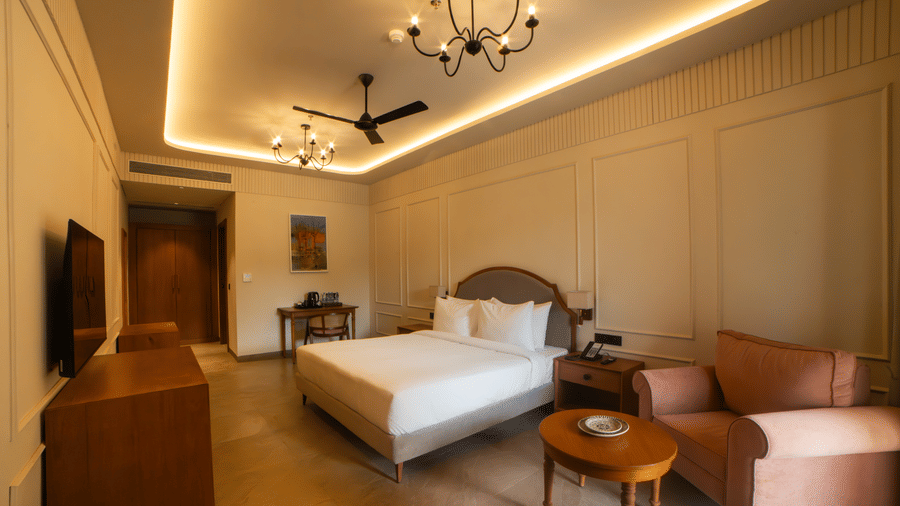 A room with a king size bed, a pink armchair with a small round table, wooden furniture, and a chandelier under a recessed lighted ceiling at Jivana Wellness Retreat, Bhopal.