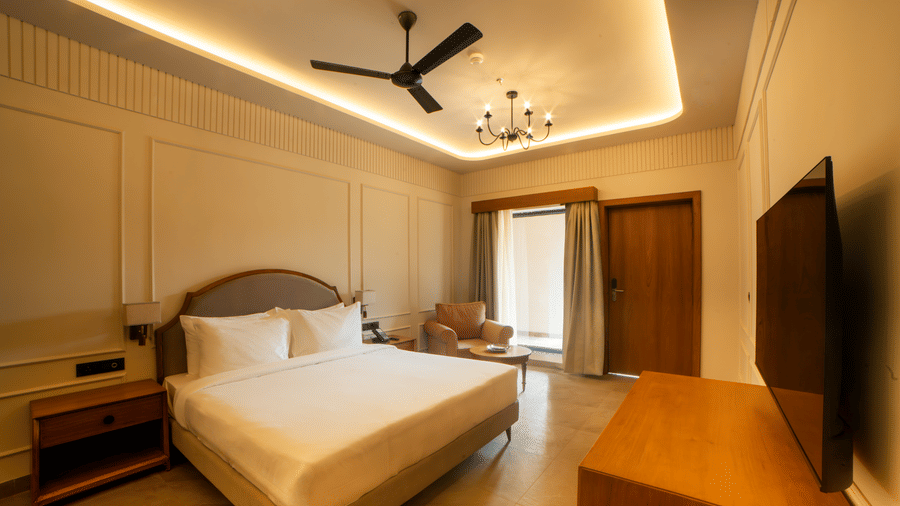 A neat room featuring a king size bed with a brown headboard, a wooden TV console, a ceiling fan, and floor-to-ceiling window at Jivana Wellness Retreat, Bhopal.