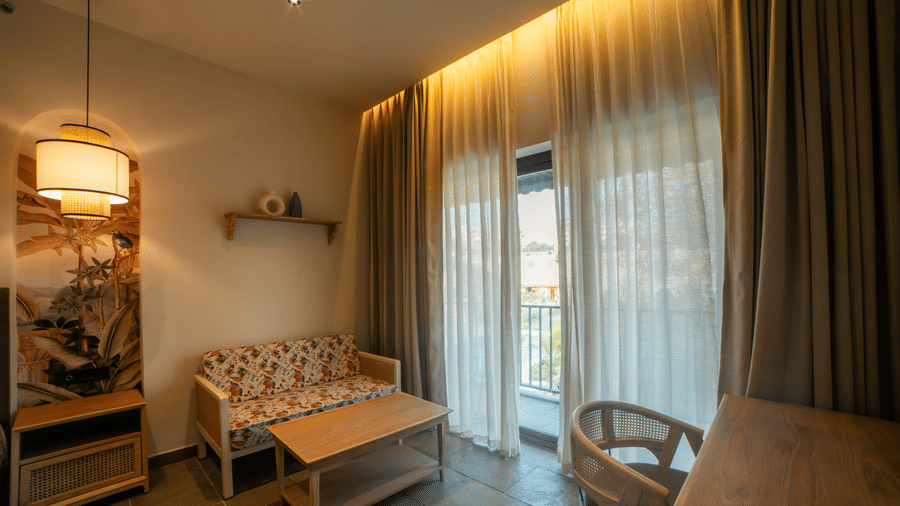 A small sitting area features a floral sofa, a light wood coffee table, a hanging lantern lamp, and heavy drapes partially covering glass doors at Jivana Wellness Retreat, Bhopal.