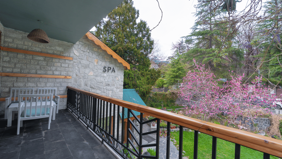 A terrace area with a stone feature wall and black railings overlooking flowering trees and the mountains - Johnson Lodge & Spa, Manali