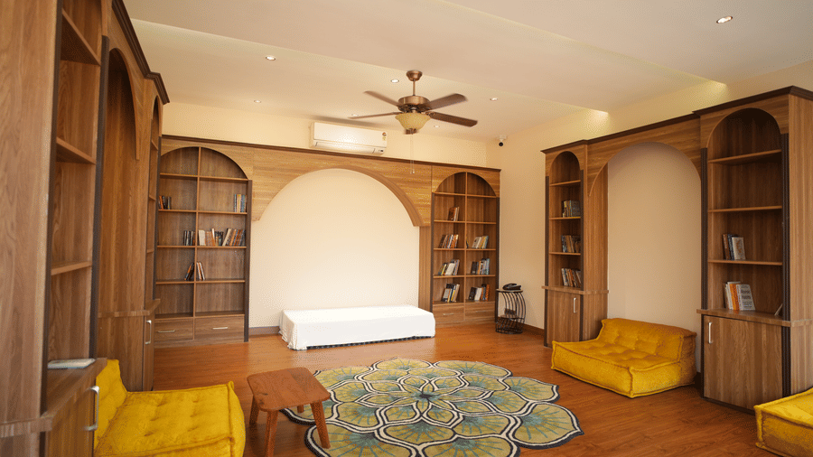 A library with built-in wooden bookshelves, a white bench, and two yellow armchairs around a green rug at Kadamb Kuteer, Vrindavan