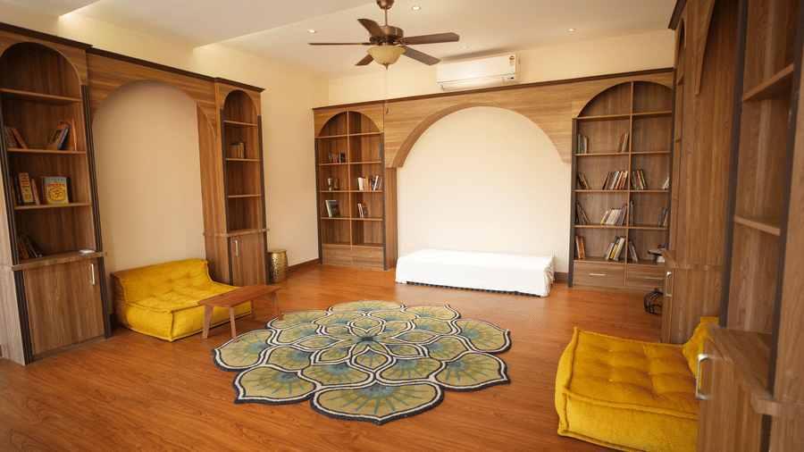 Side view of a  library with built-in wooden bookshelves, a white bench, and two yellow armchairs around a green rug at Kadamb Kuteer, Vrindavan