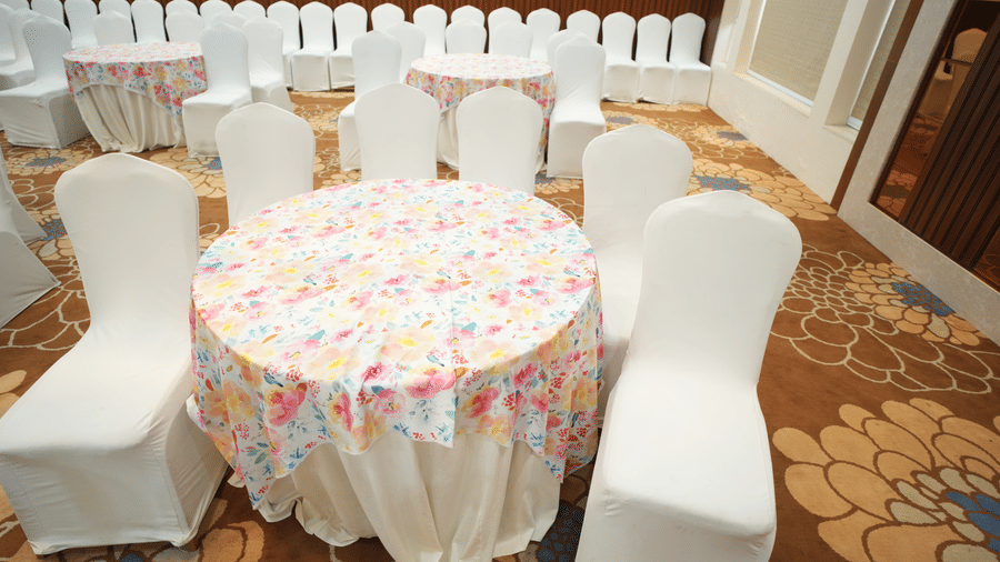 Close up of an empty banquet hall  at Kadamb Kuteer, Vrindavan with rows of white covered chairs and tables, featuring carpeted flooring and wood paneling.