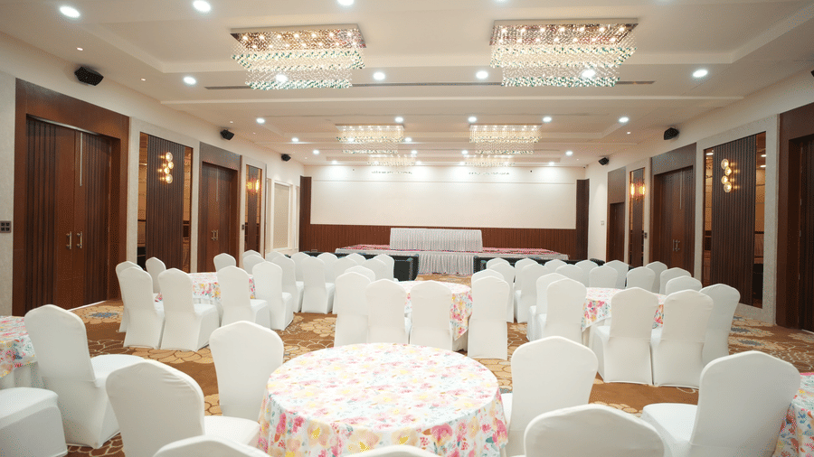 Over view of an empty banquet hall  at Kadamb Kuteer, Vrindavan with rows of white covered chairs and tables, featuring carpeted flooring and wood paneling.
