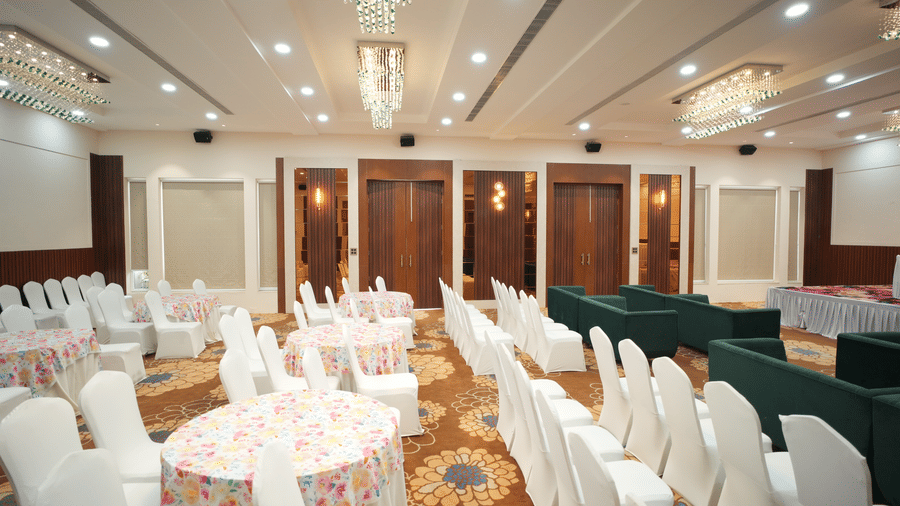 Side view of an empty banquet hall  at Kadamb Kuteer, Vrindavan with rows of white covered chairs and tables, featuring carpeted flooring and wood paneling.