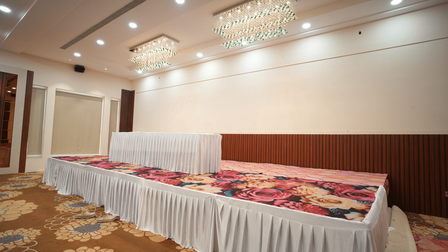 An empty banquet hall set up at Kadamb Kuteer with long, white-skirted tables and floral runners, featuring chandeliers and recessed lighting.