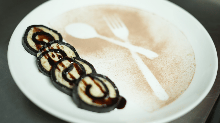 A cup of latte at Kadamb Kuteer featuring decorative chocolate art on the foam, served on a white saucer with two small biscuits on the side.