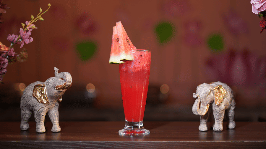 A tall glass of red layered mocktail garnished with a watermelon slice, placed next to two decorative elephant figurines at Kadamb Kuteer.