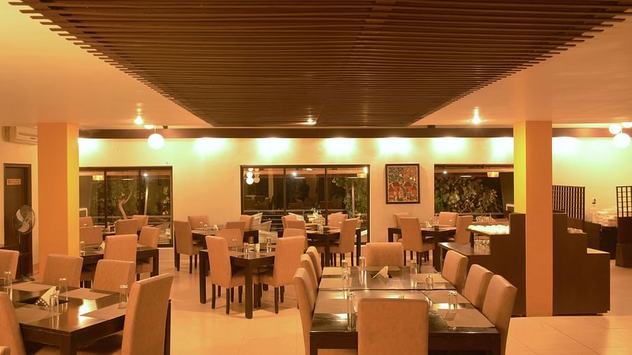 A warmly lit spacious restaurant interior with neatly arranged dining tables and chairs, a wooden slatted ceiling, and round hanging lights, reflecting a cosy dining ambience