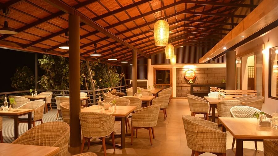 A warmly lit restaurant with an open-air feel features a wooden ceiling, tall columns, rattan chairs, and wooden tables, accentuated with elongated pendant lights and potted greenery.