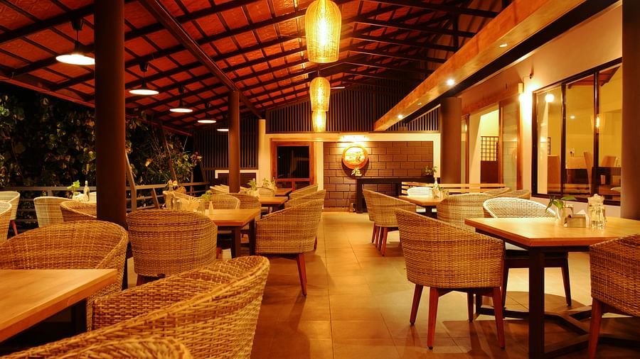 An elegant restaurant setting with woven wicker chairs, wooden tables, oriental-style pendant lights, and a high wooden ceiling, creating a warm and inviting dining atmosphere.