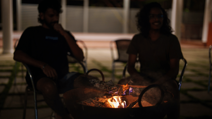 2 people sitting in metal chairs in the dark next to a burning fire inside a large metal fire pit at Kadkani River Resort, Coorg.