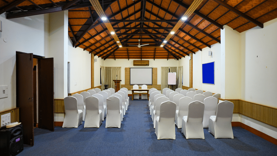 Interior view of a hall with blue carpet, white covered chairs, wooden ceiling beams, ceiling fans, and a projection screen at Kadkani River Resort, Coorg.