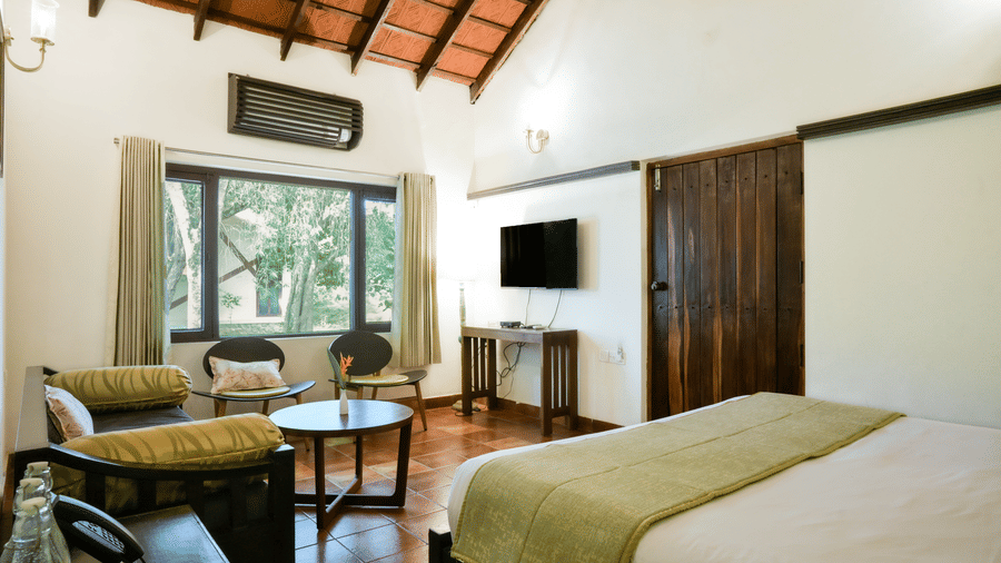 A room with a vaulted wooden ceiling features a king size bed, a wall-mounted television, an air conditioner, a round coffee table, 2 chairs, and a long daybed at Kadkani River Resort, Coorg.