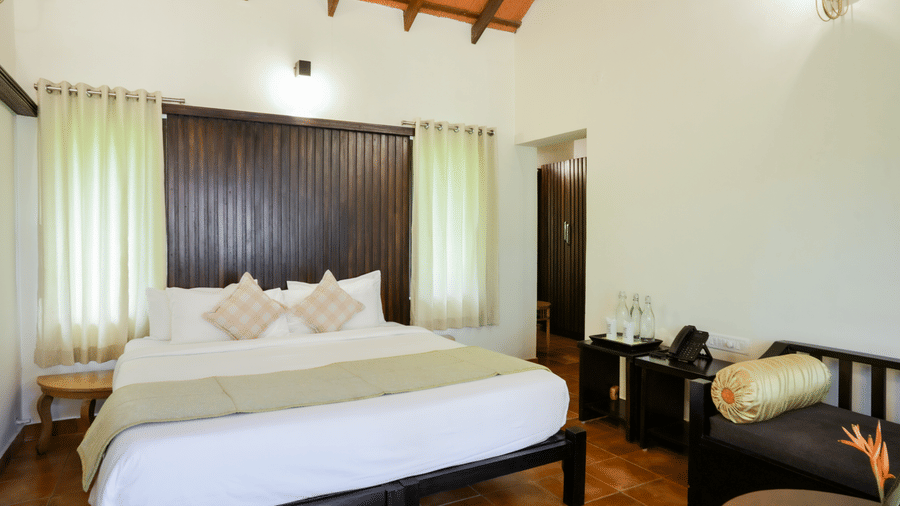 A king size bed with a light green bed runner sits facing forward, flanked by 2 curtained windows, a dark wooden panel, and a side sofa bench at Kadkani River Resort, Coorg.