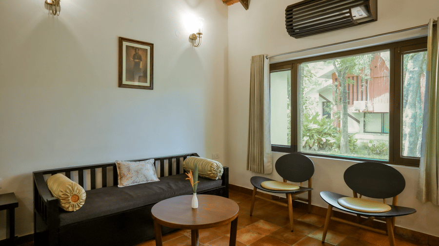 A seating area features a long dark wooden sofa bench with yellow cylindrical bolster cushions, a round wooden coffee table, 2 dark chairs, and a large window looking out at green trees at Kadkani River Resort, Coorg.